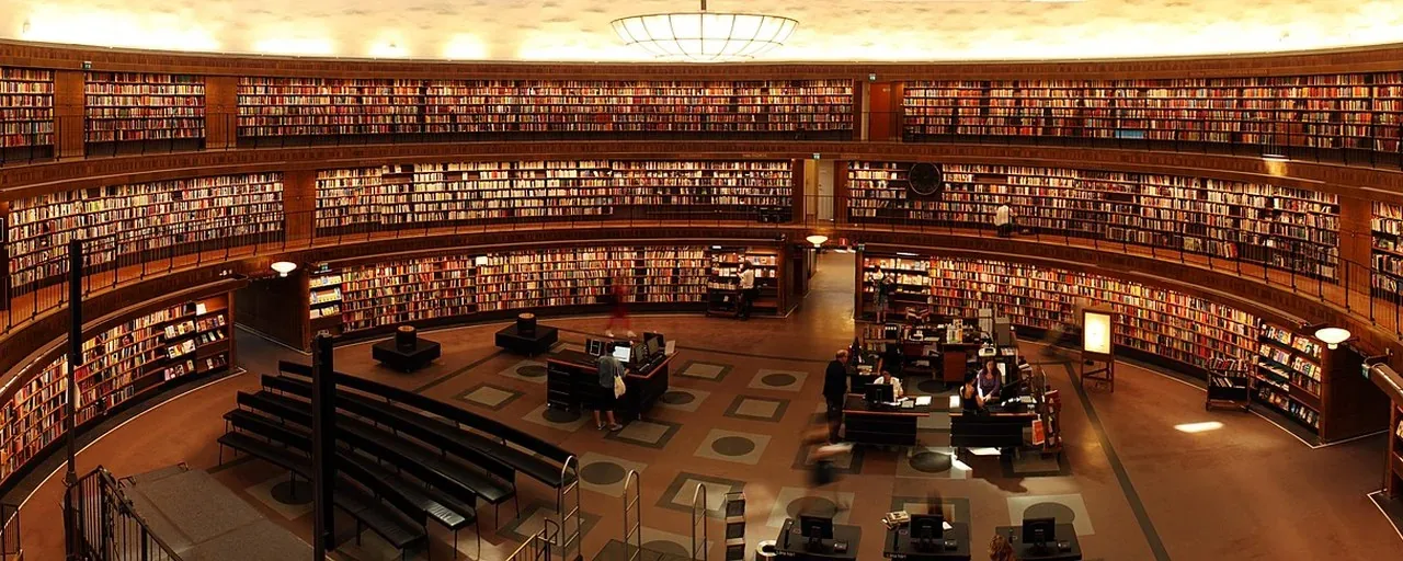 Bibliotek med bokhyllor och besökare som studerar vid läsbord på avstånd